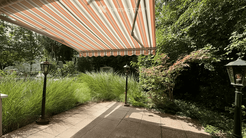 A well-maintained garden area with a striped awning, lush greenery, and decorative street lamps.