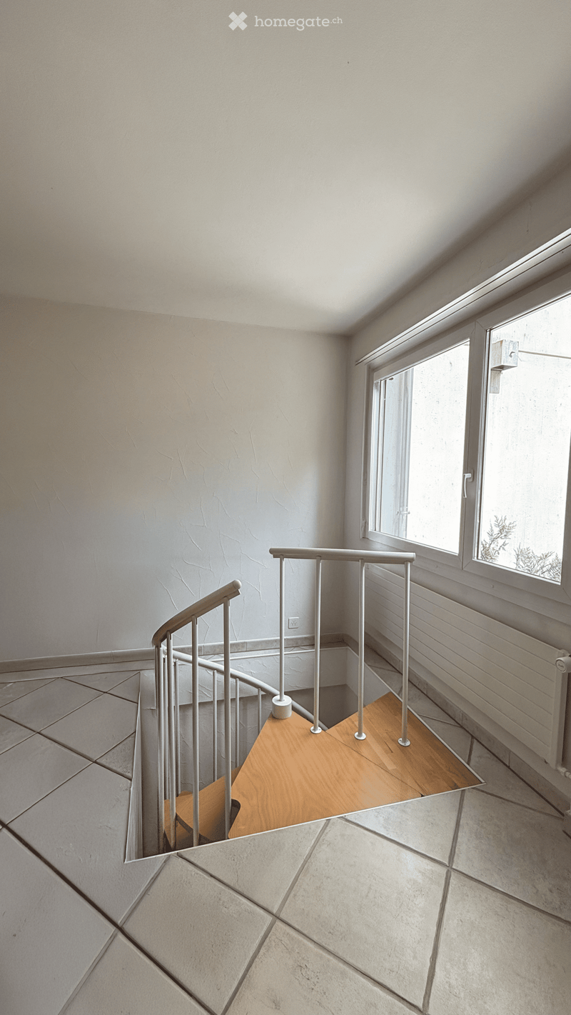 Spiral staircase with wooden steps and metal railing, light-colored walls, and a window providing natural light.