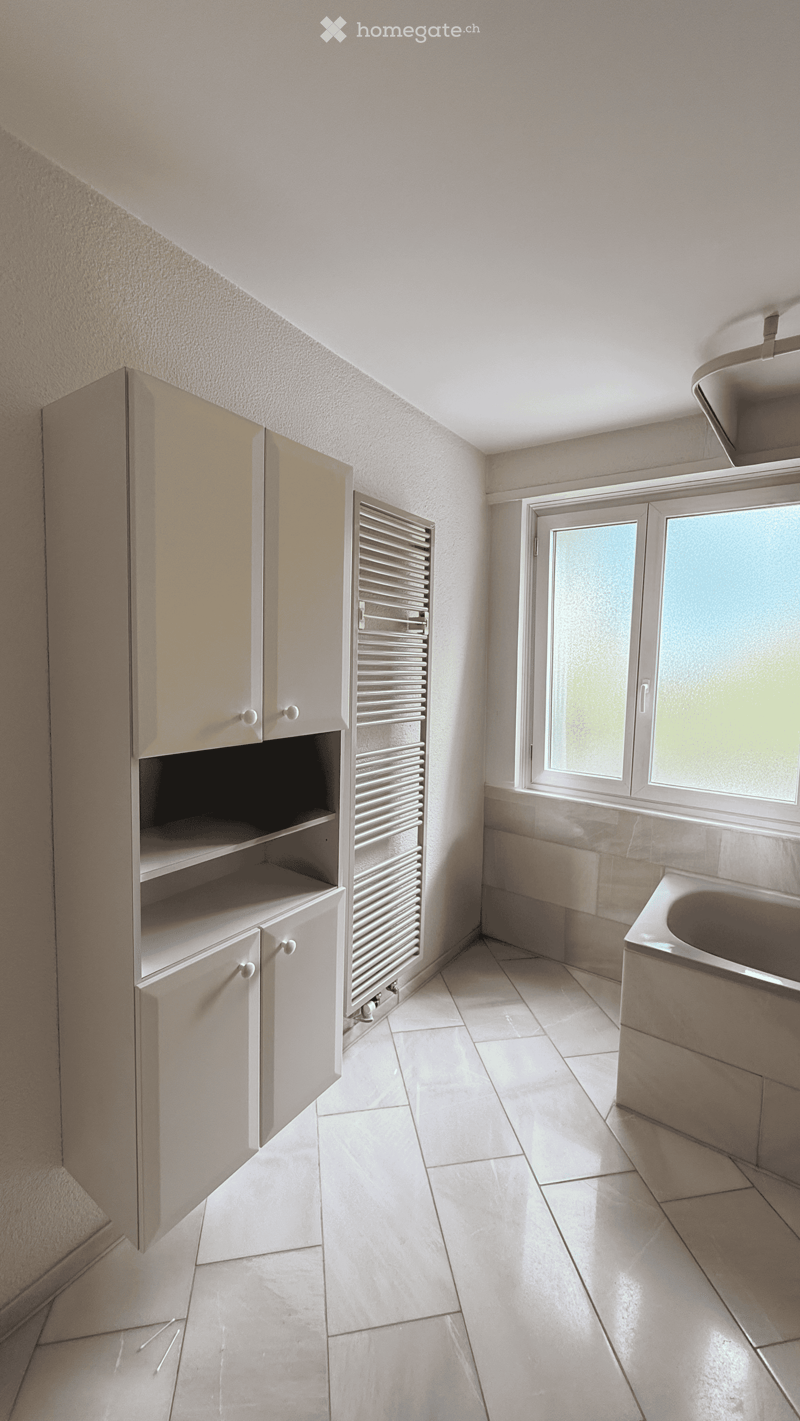 Empty bathroom with tiled floor, white cabinets, a radiator, and a window with frosted glass. No furniture or fixtures are present.