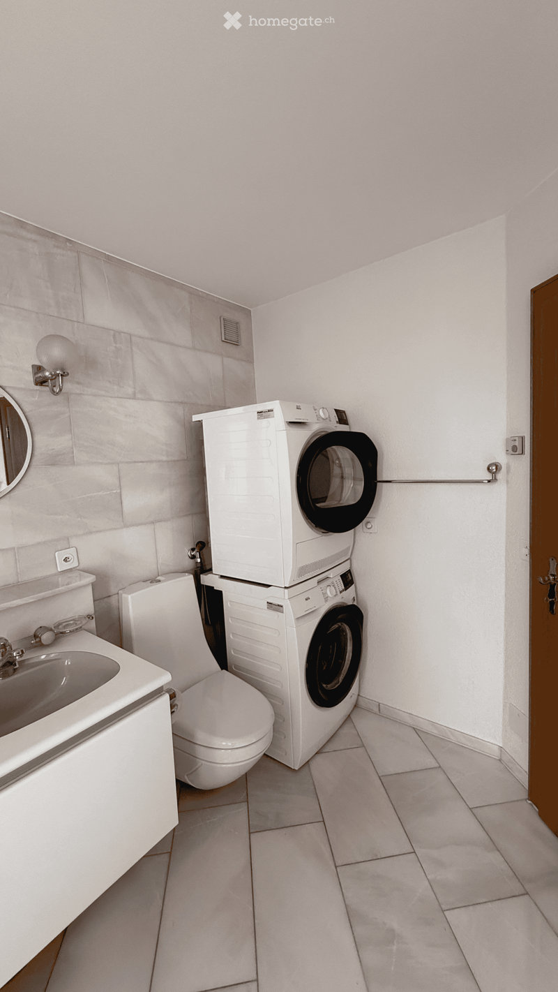 Bathroom with white tiles, a sink, toilet, washing machine, and a mirror on the wall.