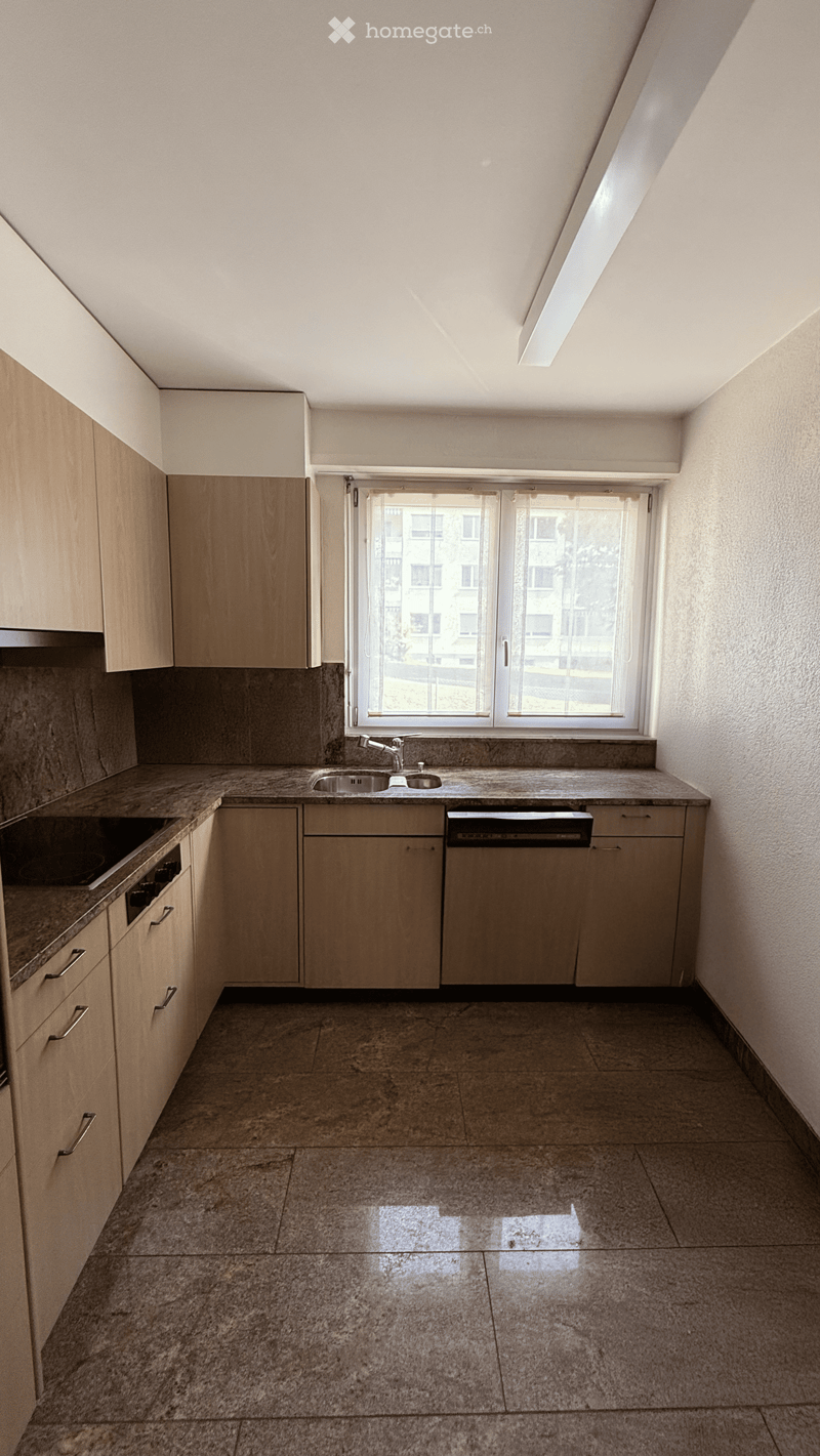 Empty kitchen with wooden cabinets, granite countertops, and a tiled floor. Features a sink, dishwasher, and a window overlooking a building.