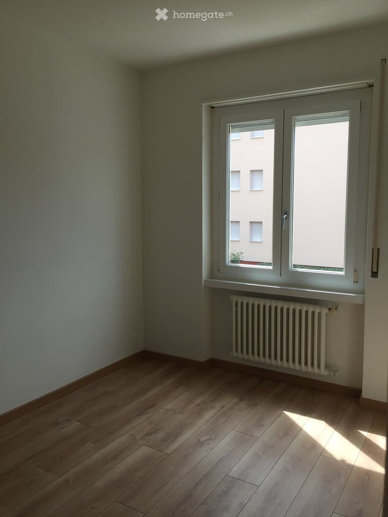 Empty room with wooden flooring, white walls, and a radiator under a large window with white frames. The room is completely unfurnished.