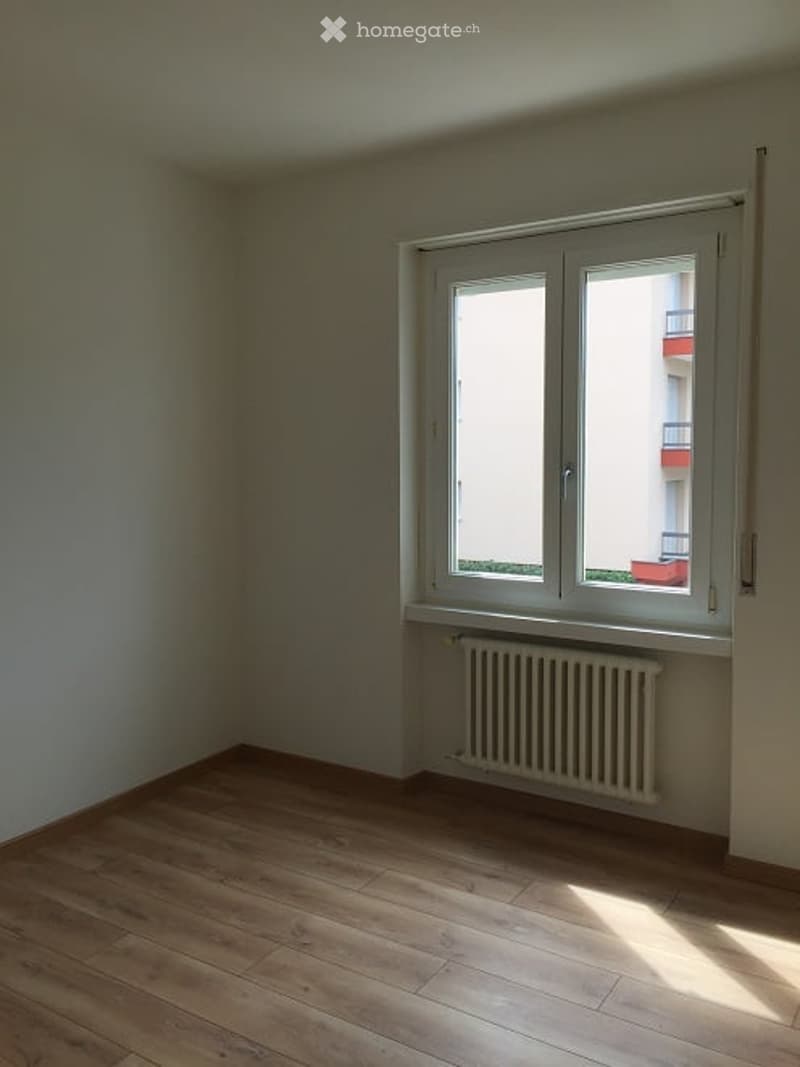 Empty room with wooden flooring, white walls, and a large window with a radiator below it. No furniture or equipment is present.