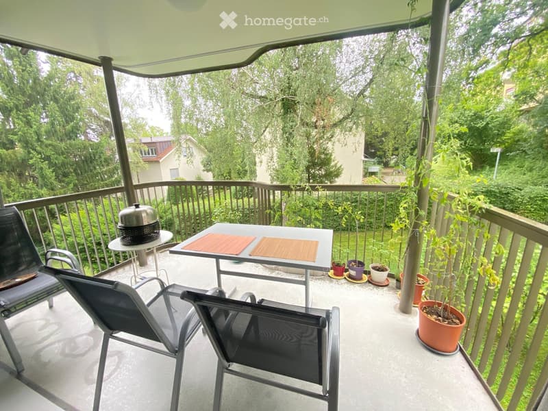 Balcony with a table, chairs, and potted plants, featuring a covered area with a railing and greenery in the background.