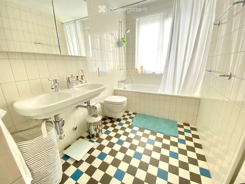 Bathroom with a sink, toilet, bathtub, and shower area. Features a checkered floor with blue, gray, and white tiles, a white tiled wall, and a mirror above the sink.