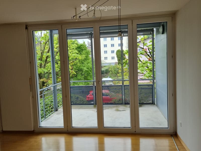 Empty room with wooden flooring, large sliding glass doors leading to a balcony, and visible greenery outside.
