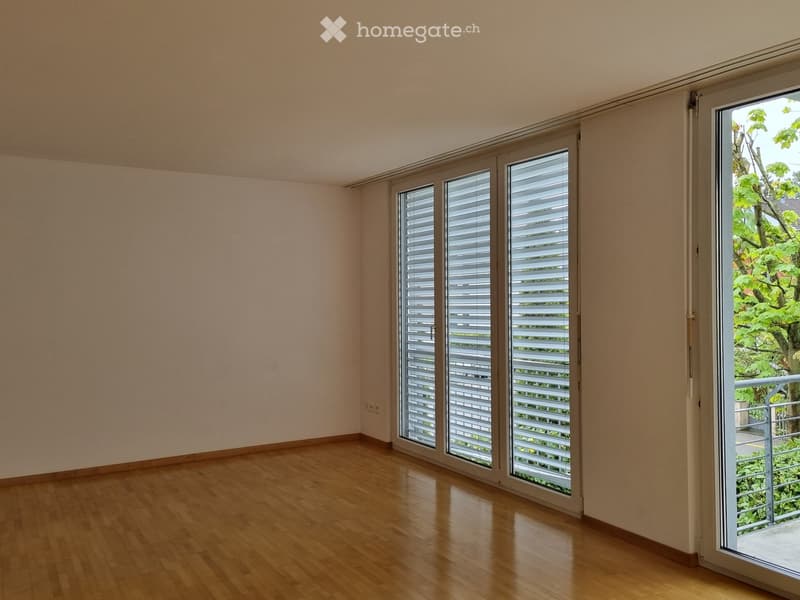 Empty room with light-colored walls and wooden flooring, featuring large windows with blinds and a sliding door leading to a balcony.