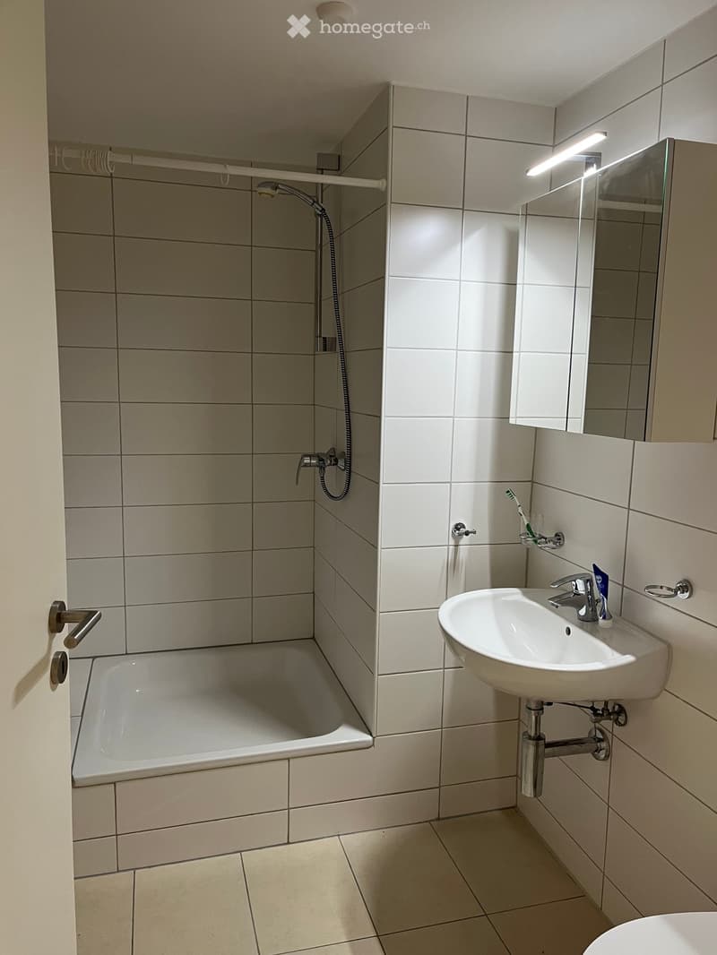Modern bathroom with a bathtub, sink, mirror, and toothbrush visible. The walls and floor are tiled in light beige.