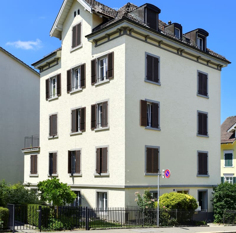 Four-story residential building with light yellow facade, multiple windows with dark shutters, and small balconies on upper floors.