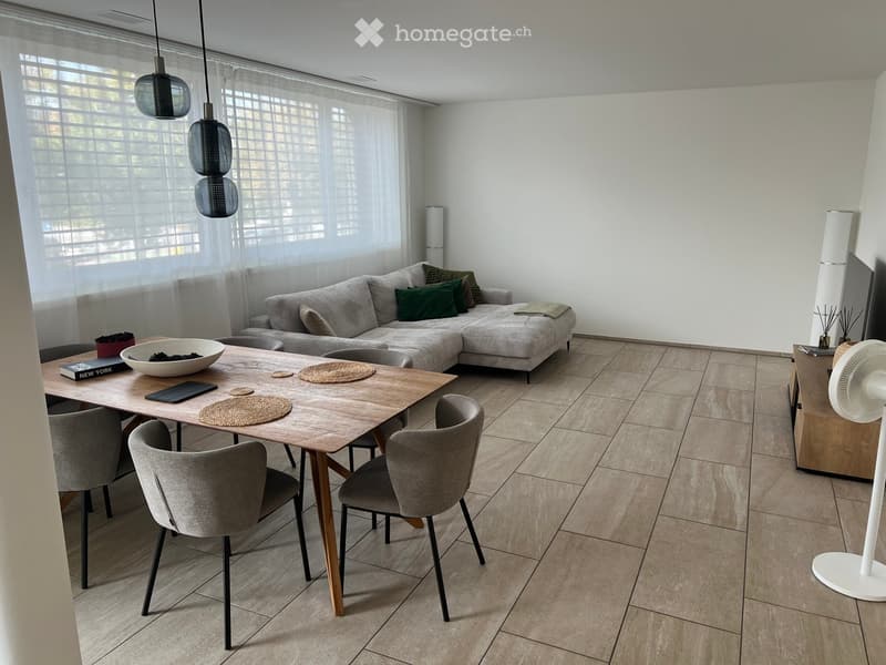 Modern living and dining area with a grey sectional sofa, wooden dining table with four chairs, decorative bowl, and a standing fan. Large windows with blinds allow natural light to fill the space.