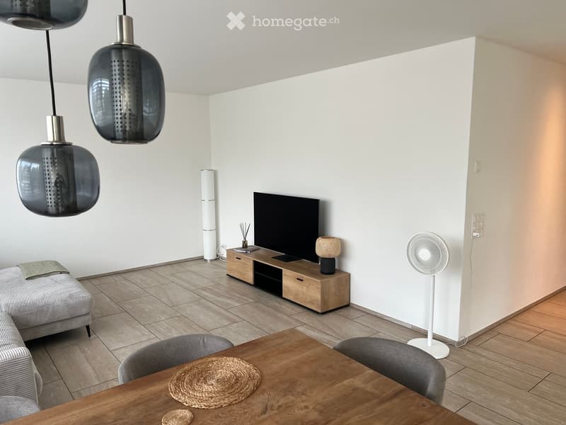Modern living room with a grey sofa, wooden coffee table, TV stand, pendant lights, and a fan. The room features a mix of wooden and tiled flooring.