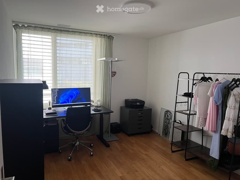 Well-lit room with wooden flooring, a desk setup with a computer and chair, a clothing rack, and a printer on a cabinet.