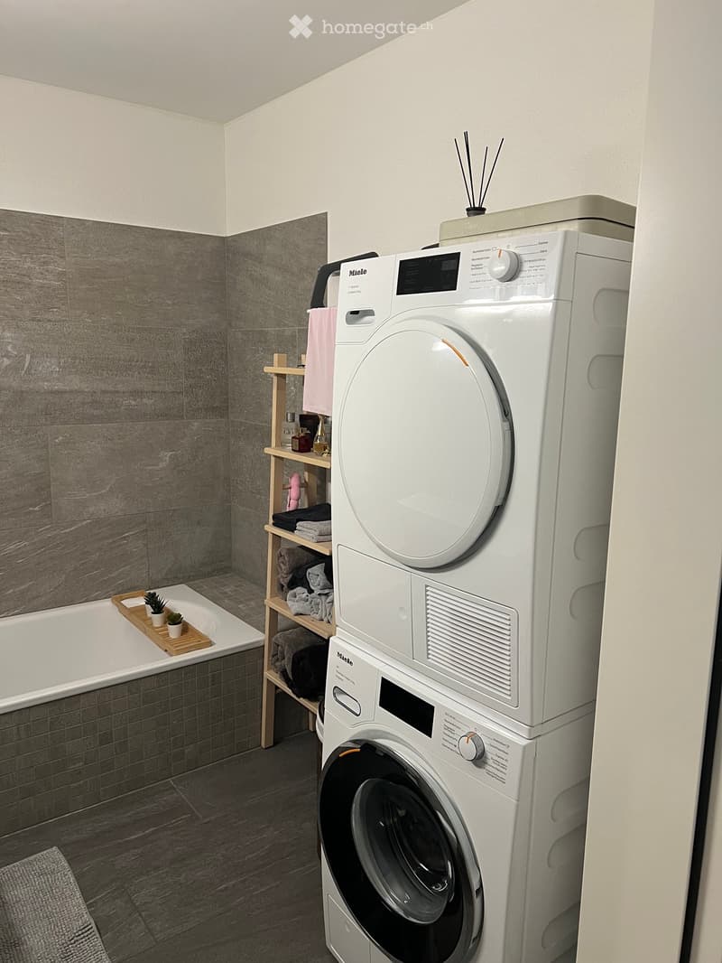 Modern bathroom with a bathtub, washing machine, wooden shelf with towels and candles, and small plants on a tray.