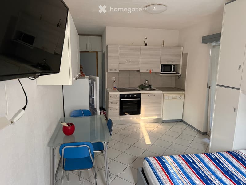 A compact studio apartment with a bed, a small dining area with a glass table and blue chairs, a kitchen area with white cabinets, an oven, a dishwasher, a microwave, and a sink.