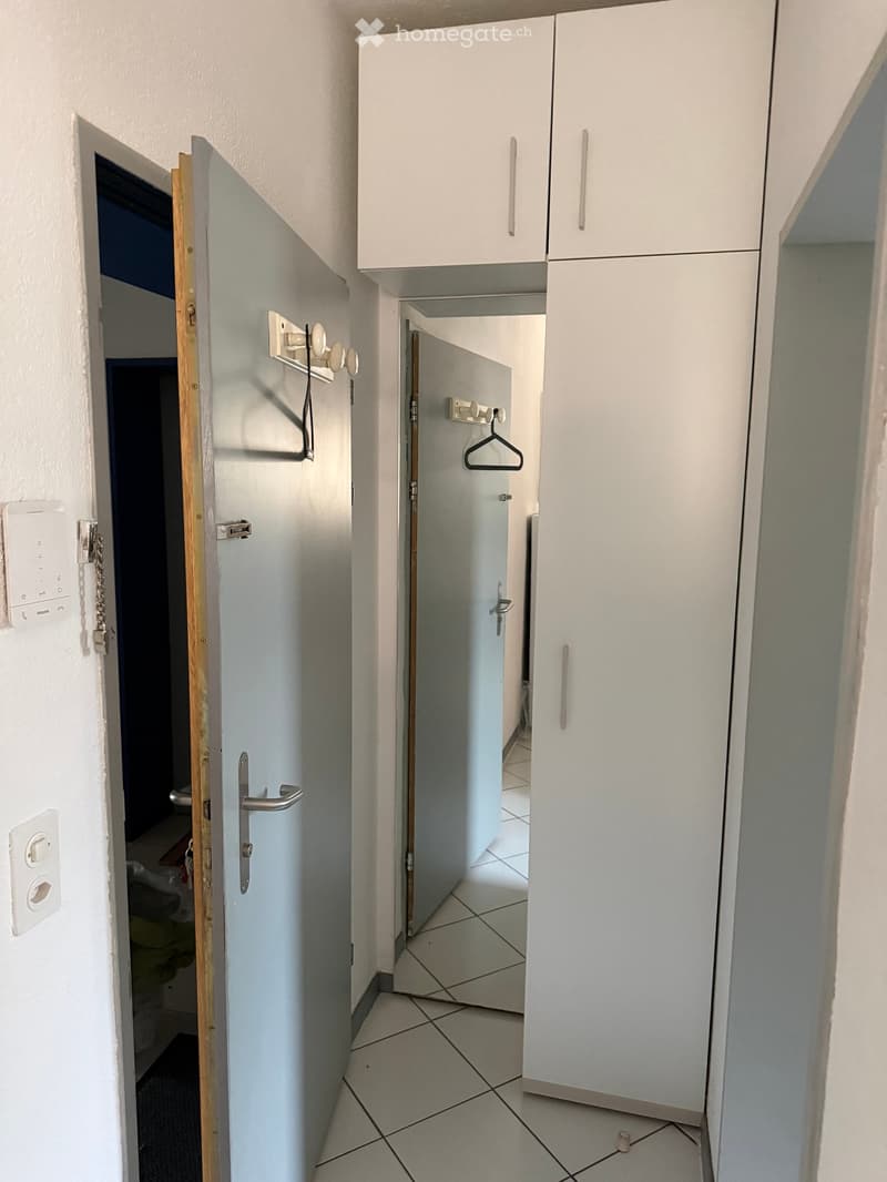 A narrow hallway with tiled flooring, white walls, and built-in white cabinets. There are coat hooks on the wall and a partially open door leading to another room.