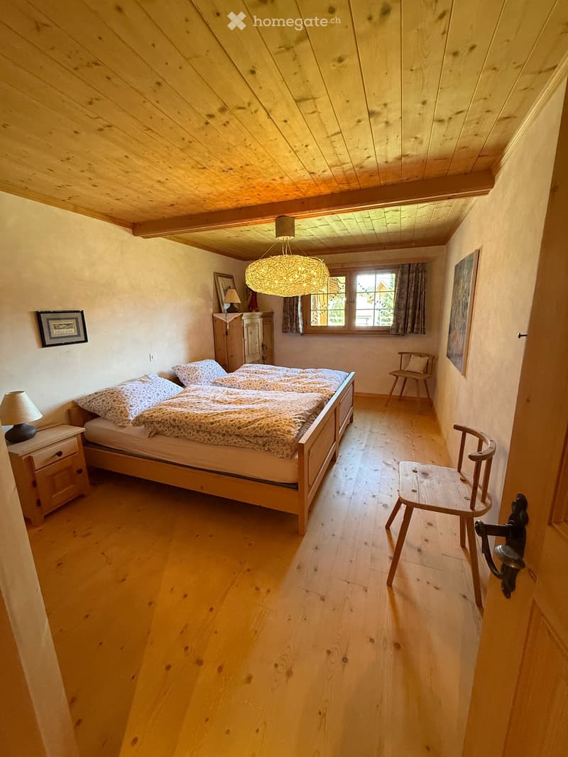Cozy bedroom with wooden floors, slanted wooden ceiling, large window with curtains, bed with patterned bedding, nightstand with lamp, wooden chair, and decorative lighting fixture.