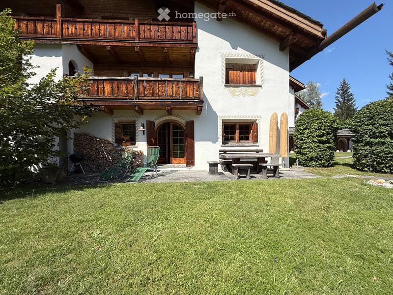 Two-story white house with wooden balconies, large windows, and a well-maintained lawn. Outdoor seating area includes a picnic table, benches, and two green chairs.