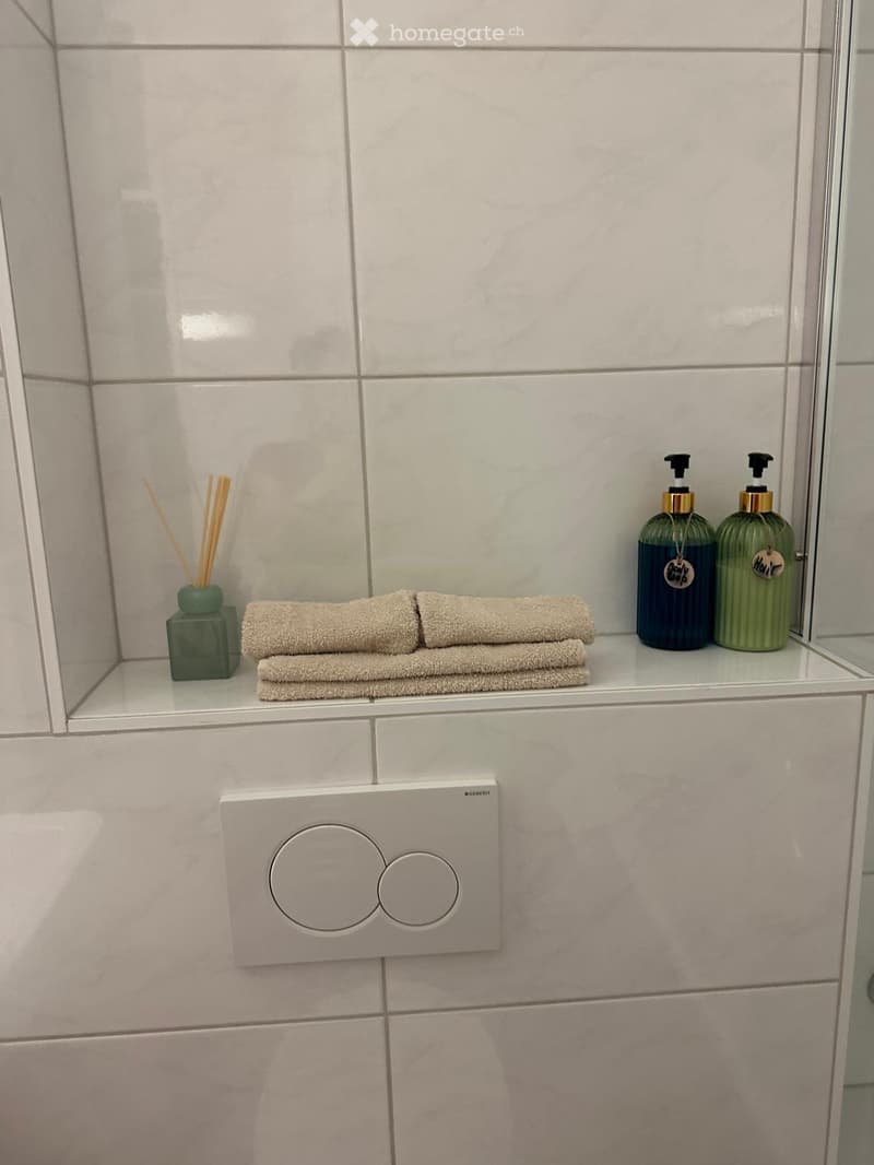 Bathroom with light grey tiles, a white shelf holding folded towels, two soap dispensers, and a decorative diffuser.