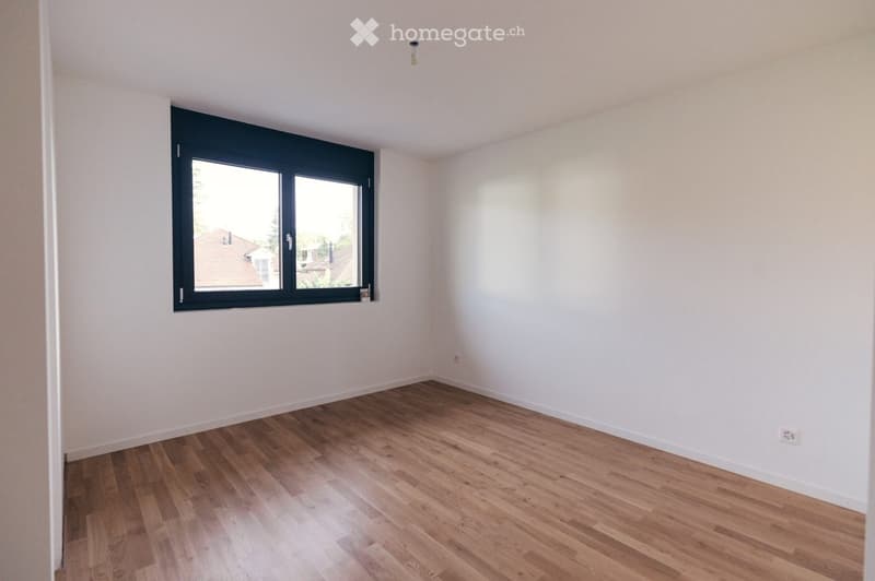 Empty room with light wooden flooring and white walls, featuring a large window with black frame and a visible outdoor view.