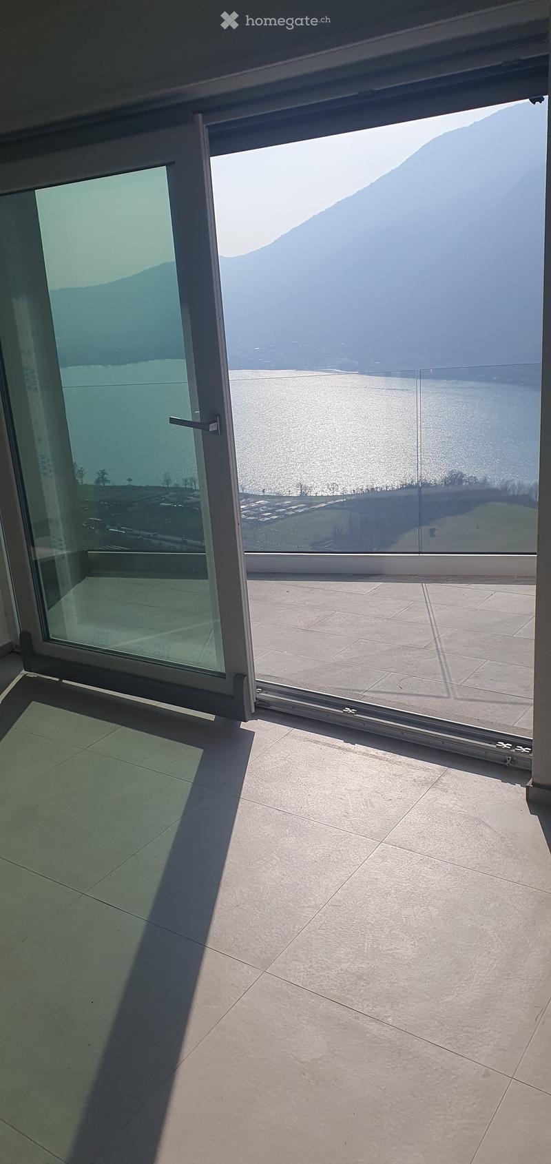 Balcony / Terrace. Large balcony with expansive view of a lake and mountains, featuring modern glass doors and tiled flooring.