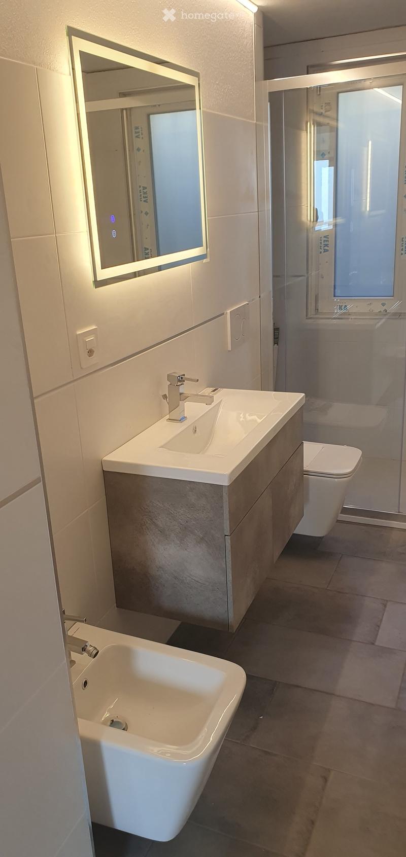 Bathroom. Modern bathroom with a white sink, chrome faucet, illuminated mirror, toilet, and bathtub. The walls are tiled in light gray, and the floor is dark gray.