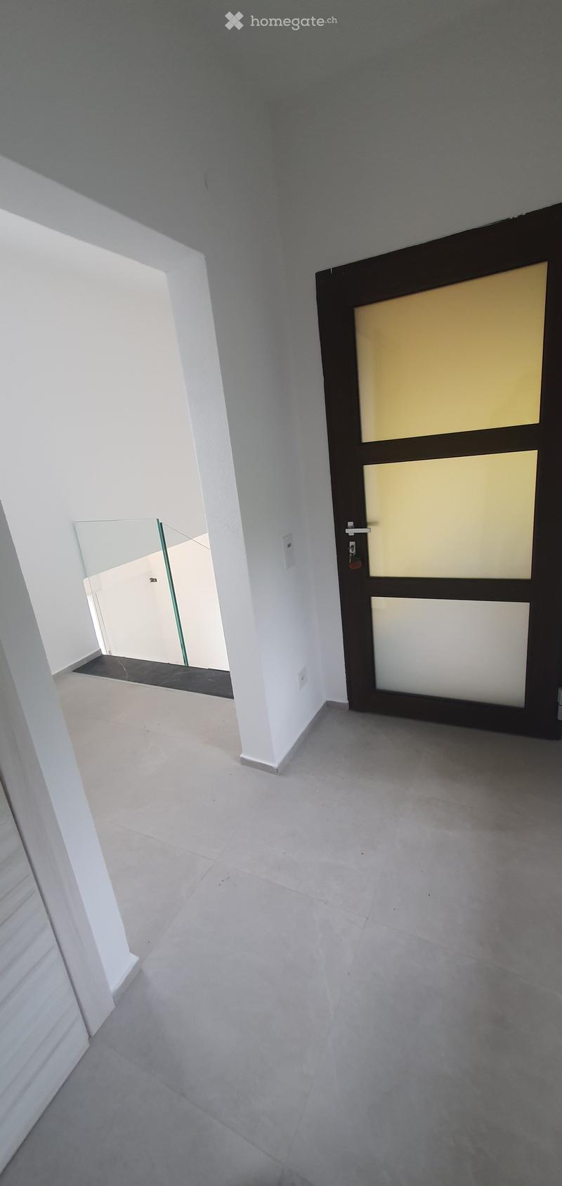 Corridor / Hallway. Empty hallway with white walls, grey tiled floor, and a closed door with glass panels on the right side.