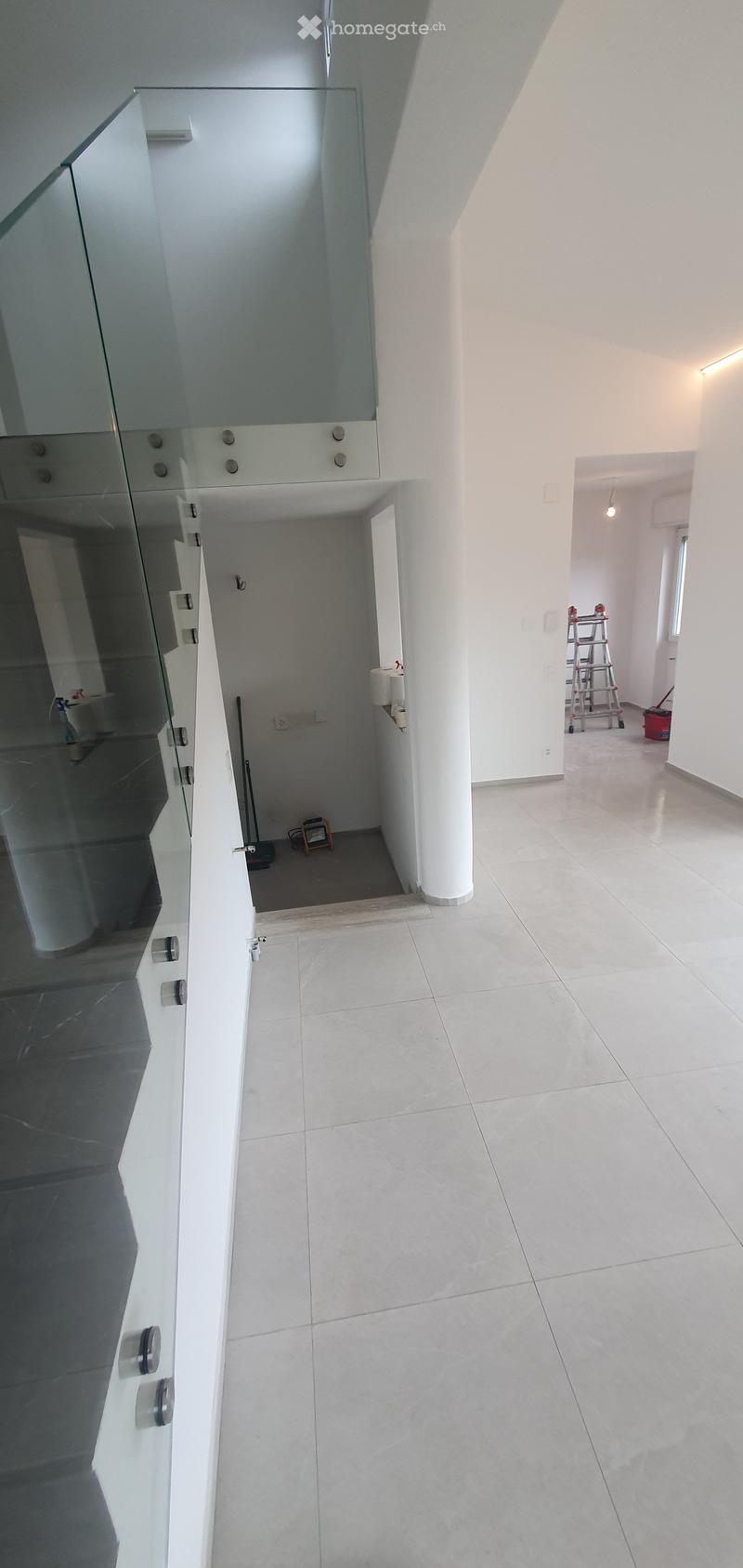 Corridor / Hallway. Empty hallway with white walls, tiled floor, and a glass door on the left side. A white column is visible in the center, and a small room with a ladder and cleaning supplies is visible at the end.