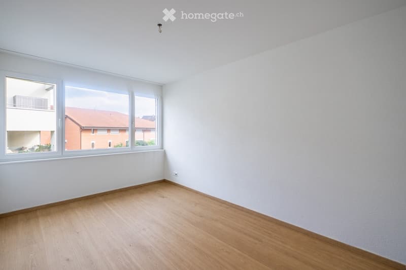 Empty room with light wooden flooring, white walls, and large windows offering a view of neighboring buildings.