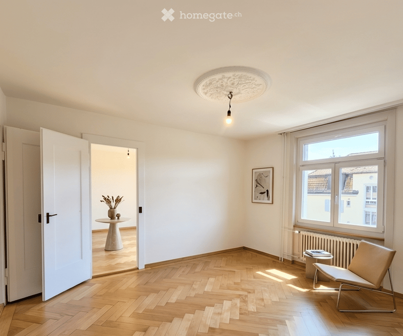 Bright, unfurnished room with wooden flooring, large windows, a single chair, a small table with a vase, and a picture frame on the wall.