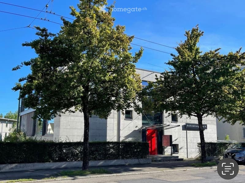 Modern two-story building with a concrete facade, large windows, and a red entrance door. Surrounded by trees and greenery.