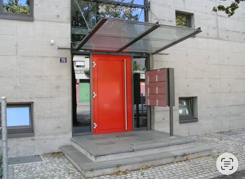 Modern building entrance with a bright red door, glass canopy, and steps leading up to the entrance.