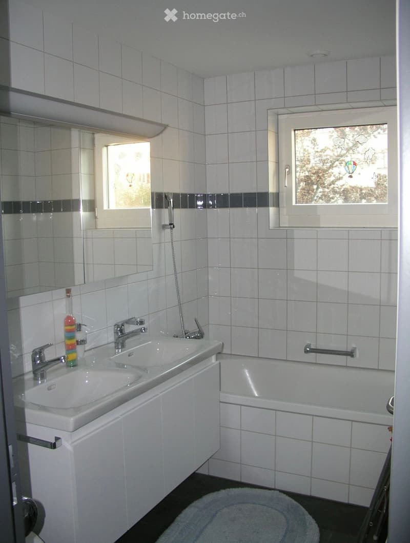 Modern bathroom with white tiles, double sink, bathtub, and large windows offering a view of the outside.