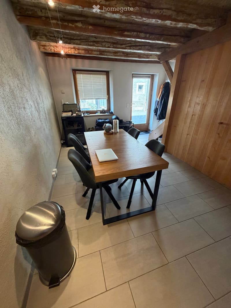 Dining area with a wooden table, four chairs, a laptop, a small cabinet, and a trash can. The room has exposed wooden beams on the ceiling and a window with blinds.