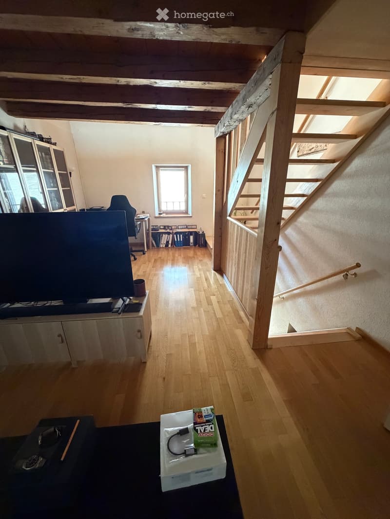 Bright living room with wooden floors, exposed wooden beams, TV on stand, chair, small table, and cabinet. Stairs lead to another level.
