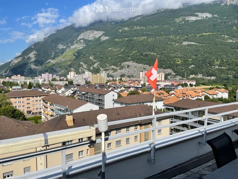 Balcony overlooking a town with red-tiled roofs and a mountainous backdrop, featuring a Swiss flag and a weather station.