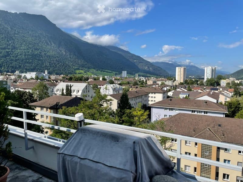 Balcony with a scenic view of a cityscape and mountains, featuring a covered object and a small potted plant.