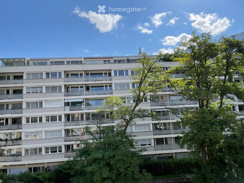 Multi-story residential building with numerous windows and balconies, surrounded by trees and greenery.