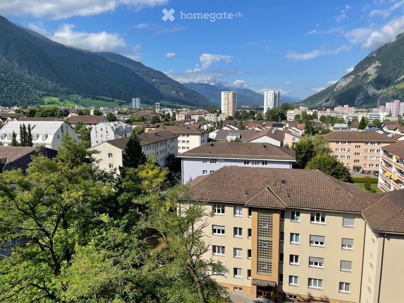 A panoramic view of a town nestled in a valley with surrounding mountains, featuring multiple residential and commercial buildings with varied architectural styles and roof types.