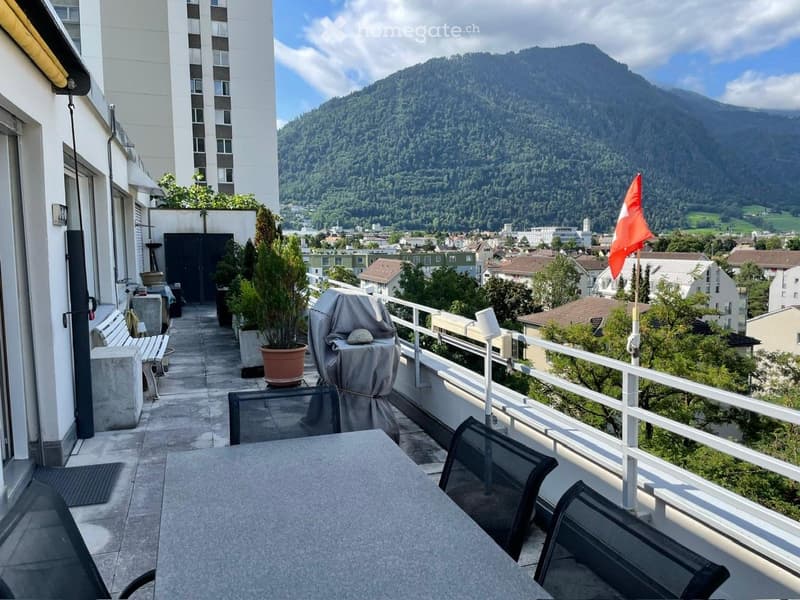 Balcony with a scenic mountain view, featuring a table, chairs, a bench, potted plants, and a covered grill.