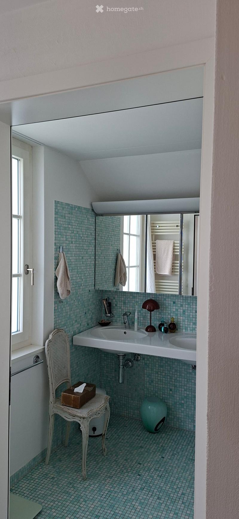 A small, modern bathroom with a double sink, a large mirror, a wicker chair with a box on it, a vase, and various toiletries on the sink counter.