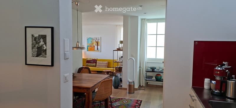 Open-plan living and dining area with a yellow sofa, wooden dining table, wicker chairs, a coffee maker, and a sleek speaker on the floor. The space features modern decor, including framed artwork and a colorful rug.