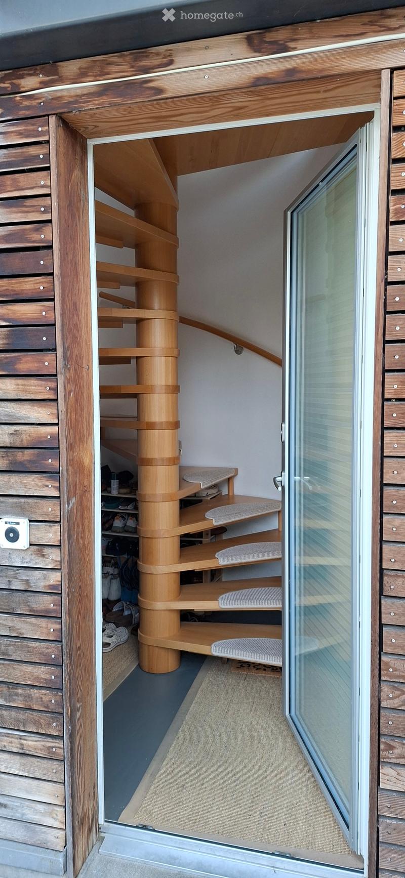 Spiral staircase with wooden steps and shelves, leading to a small storage area with shoes and bags. The hallway features wooden walls and a glass door leading outside.