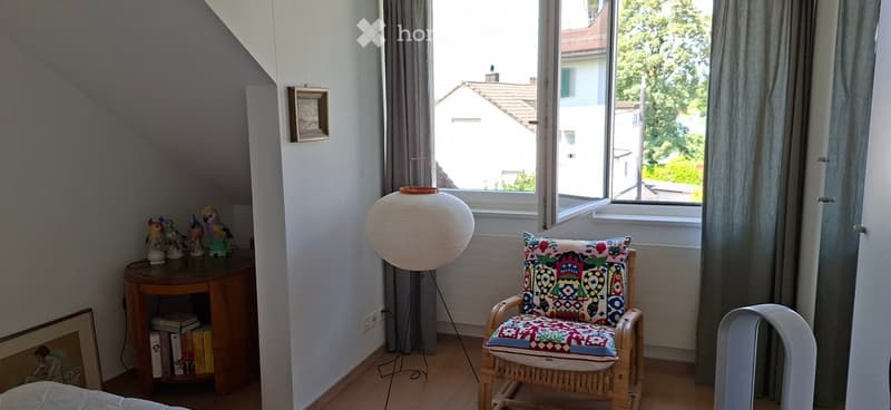 A cozy, well-lit room with wooden flooring, a round wooden table with books and decorative ornaments, a wicker chair with a colorful cushion, and a large white floor lamp near the window.