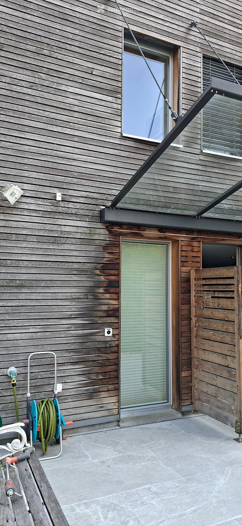 Wooden exterior wall with large windows and a glass roof extension, featuring a garden hose and a white chair on a stone patio.