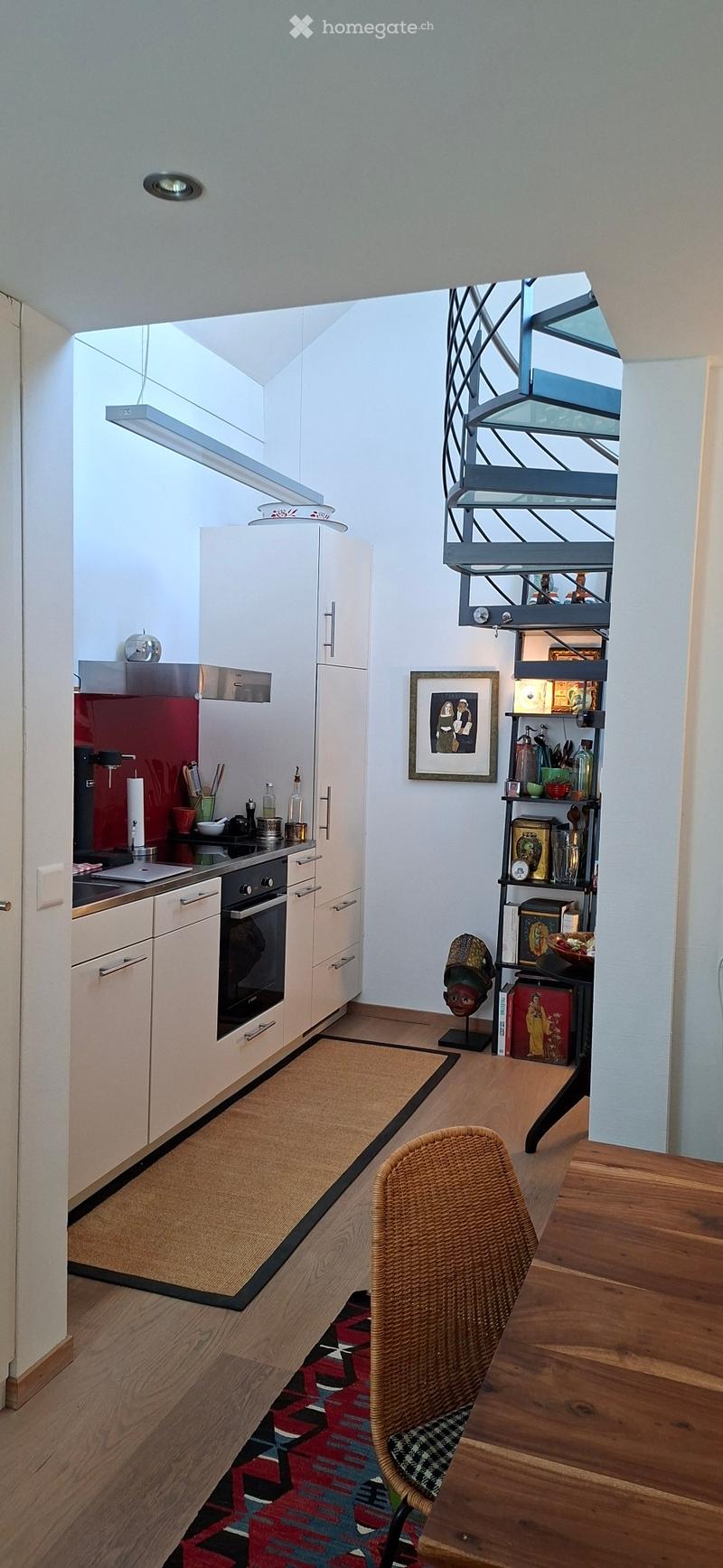 Modern kitchen with white cabinets, stainless steel appliances, wooden flooring, and a small seating area with a wicker chair.
