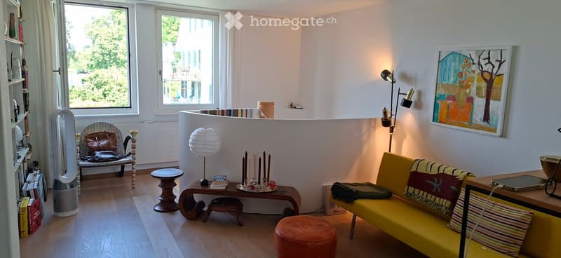 Bright living room with wooden flooring, large windows, a yellow sofa with colorful cushions, a wooden chair with a patterned cushion, a small round table, a floor lamp, and a bookshelf with books and decorative items.