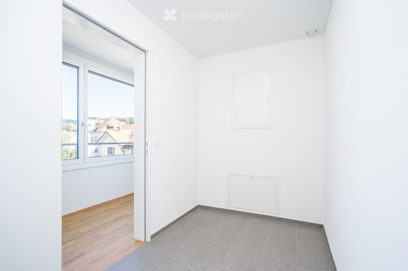 Empty room with white walls, wooden flooring, and large windows offering a view of the outside. No furniture or equipment is present.