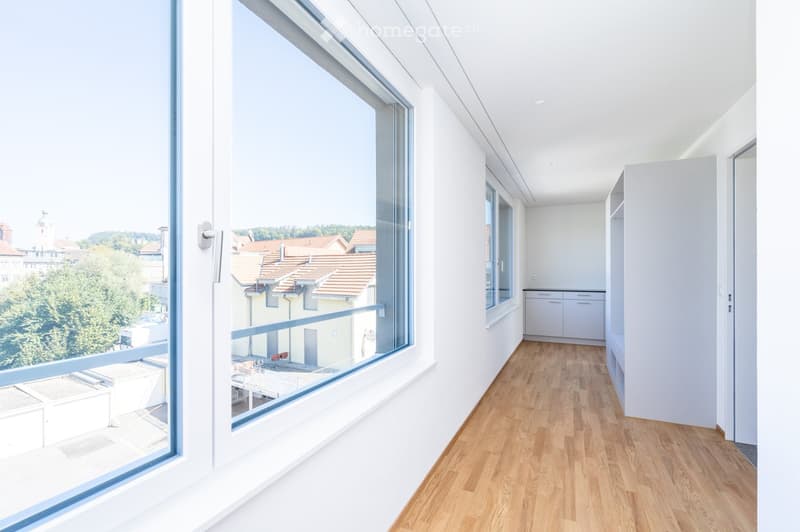 A modern, minimalist hallway with light wooden flooring, white walls, and large windows offering a view of the cityscape and surrounding greenery. A small cabinet is visible on the right side.