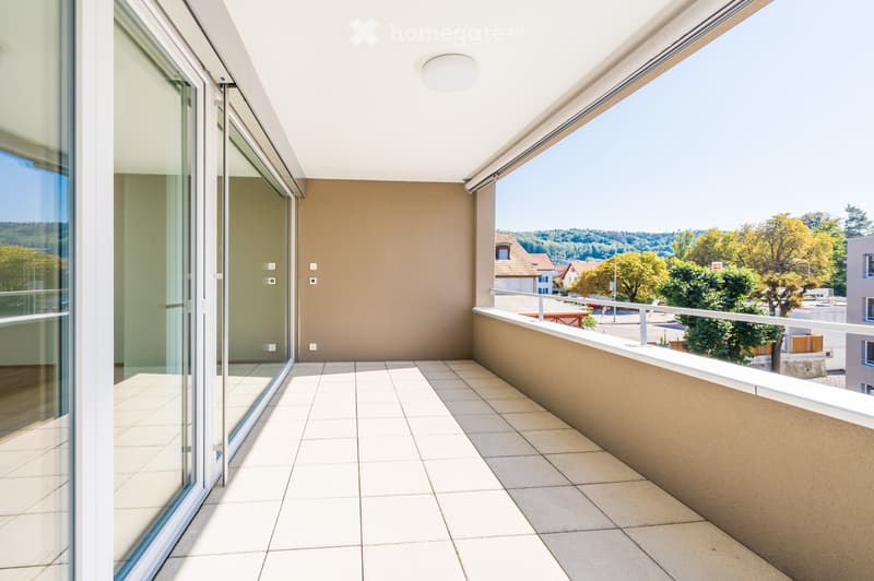 Modern balcony with tiled flooring, large glass sliding doors, white ceiling light, and a view of a green landscape with trees and houses.