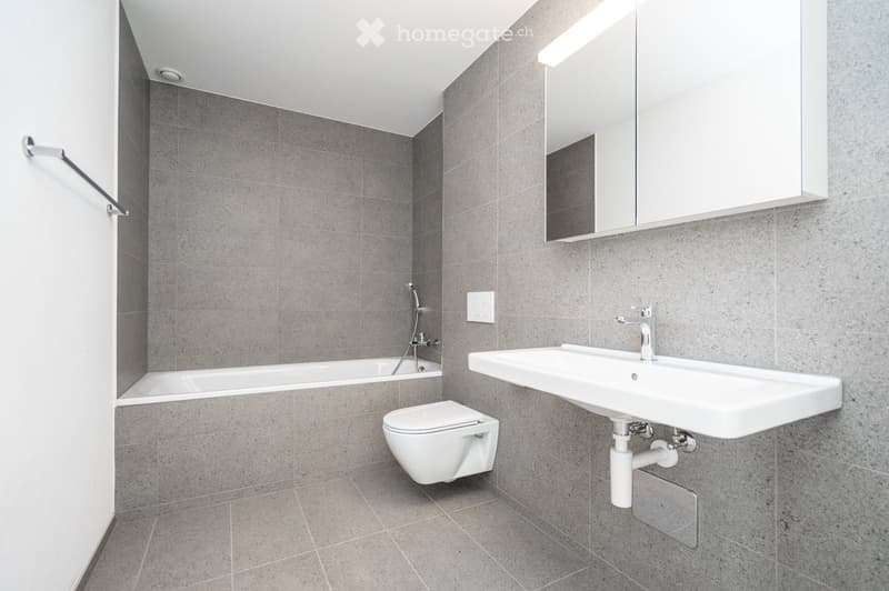 Modern bathroom with grey tiled walls and floor, featuring a bathtub, toilet, sink, and mirror.